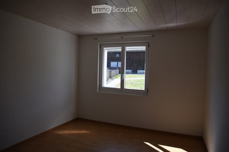Empty room with wooden flooring, white walls, and a large window offering a view of a grassy area and wooden fence.