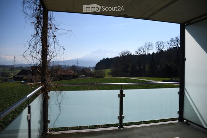 Balcony with glass railing, overlooking a green field, trees, and distant mountains.