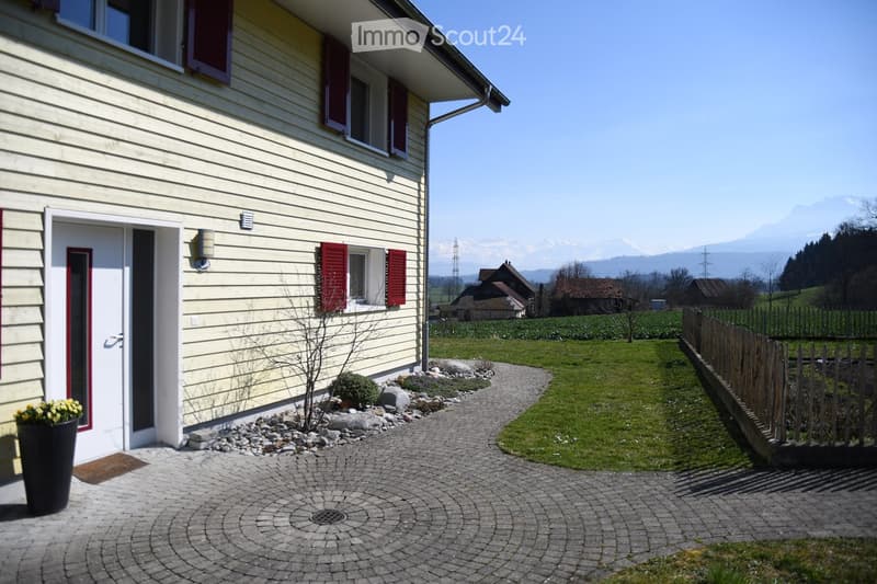 Front garden area with a paved pathway leading to the house entrance, small plants, a wooden fence, and a scenic mountain view in the background.