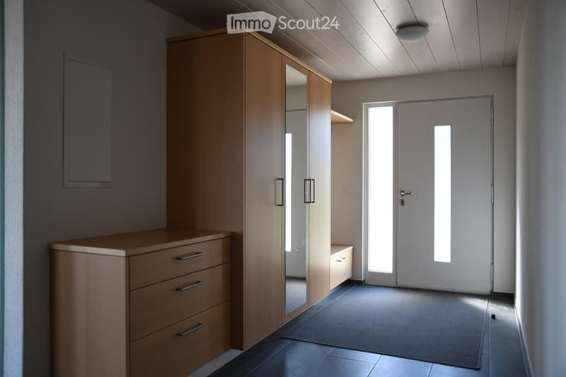 Empty room with wooden furniture including a tall wardrobe with mirrored doors, a chest of drawers, and a small bench. The room has a tiled floor and a large white door with glass panels.