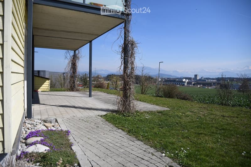 A paved pathway leads to a covered balcony area with a view of a grassy lawn, distant fields, and mountains. Small purple flowers are planted along the pathway.