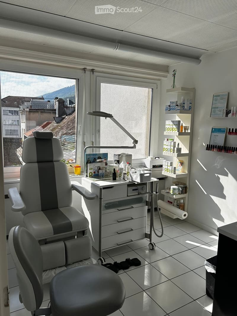 A modern, well-lit room with a large window offering a view of rooftops and mountains. The room features a professional chair, a table with various items, shelves with products, and a lamp.