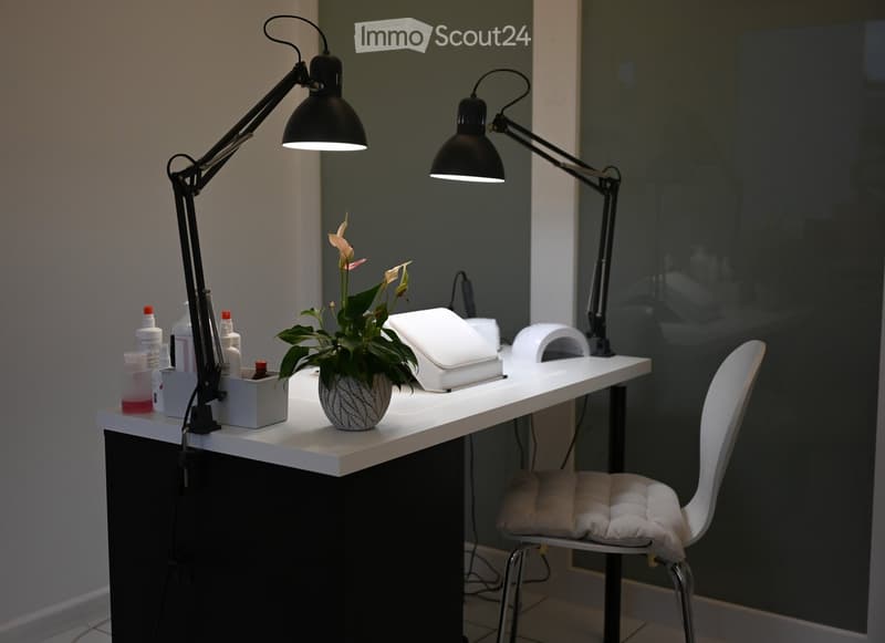 A well-lit, modern nail station with a white table, a white cushioned chair, a potted plant, two adjustable lamps, and various nail care products and tools.