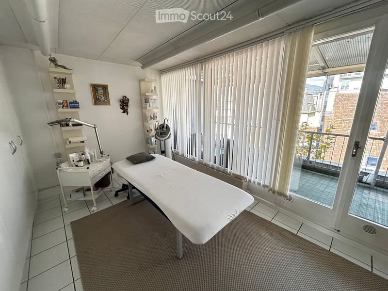A well-lit, modern treatment room with a white treatment table, a small cabinet, shelves with various items, a lamp, and a mirror. The room has large windows with vertical blinds and a view of the outside.