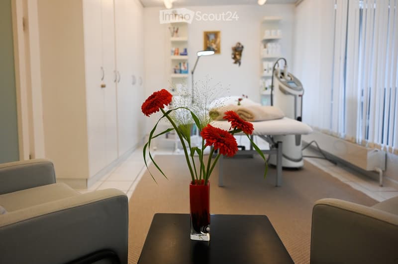 A modern, well-lit room with a sleek design, featuring a grey sofa, a small black table with a vase of red flowers, a treatment table with towels, and a cabinet with shelves.