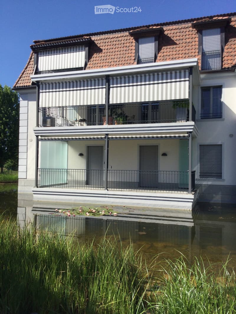 Two-story house with a raised ground floor, surrounded by water, featuring balconies, striped awnings, and a small pond with lily pads in front.