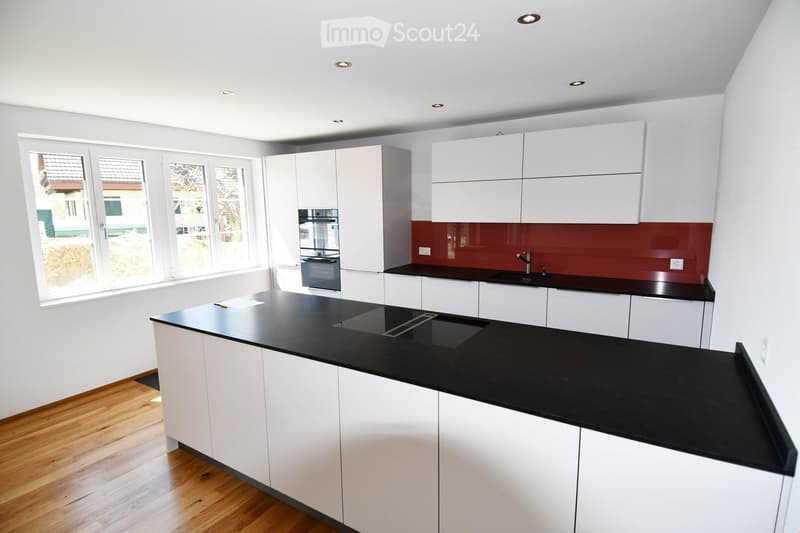 Modern kitchen with white cabinets, black countertops, and a red backsplash. Features include a central island, built-in oven, and large windows offering a view of the exterior.