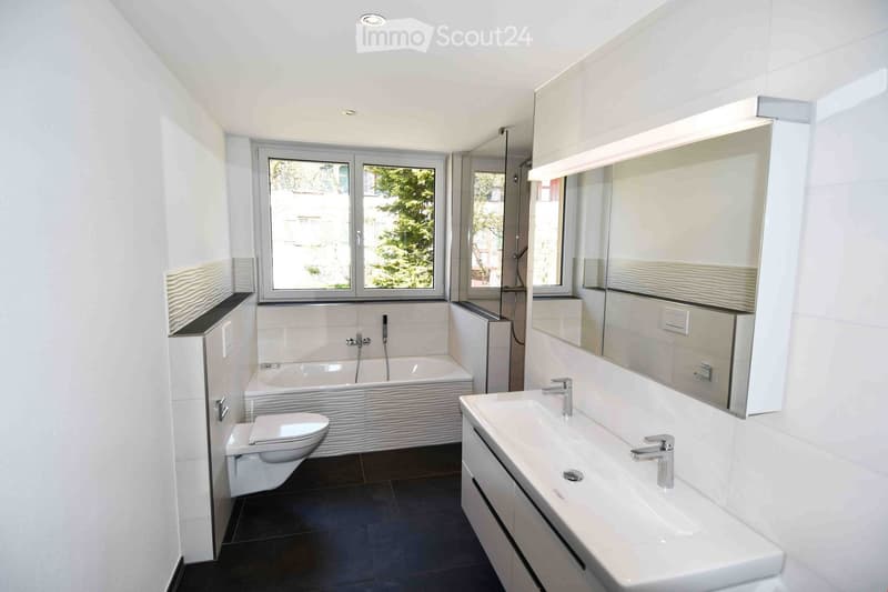 Modern bathroom with white tiles, a bathtub, toilet, sink, large mirror, and built-in cabinets.