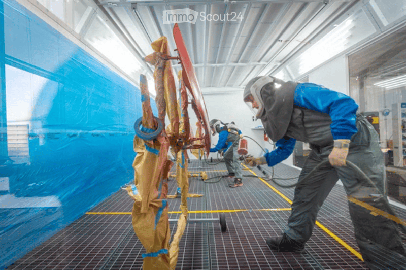 Two workers in protective gear are spray painting a large red object in a well-lit industrial space, using spray guns and protective masks.