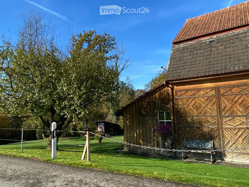 A wooden barn with a steep roof and large wooden doors, surrounded by a grassy area with trees and a bench near the entrance.