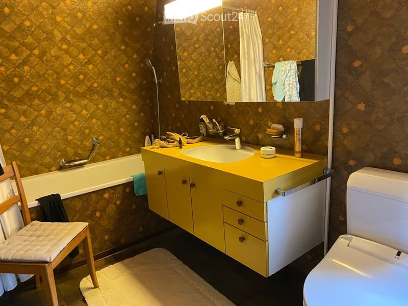 Bathroom with yellow vanity, sink, bathtub, toilet, and a wooden chair with a cushion. The walls have patterned wallpaper, and towels are hanging on racks.