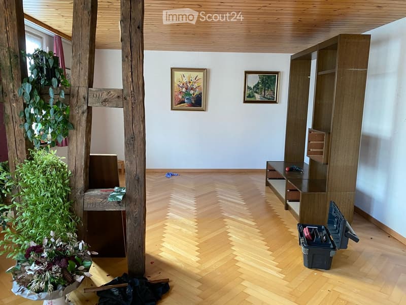 Empty room with wooden herringbone flooring, white walls, exposed wooden beams, and a window with a plant on the sill. There are some scattered items like a toolbox, a cloth, and a small wooden bench.