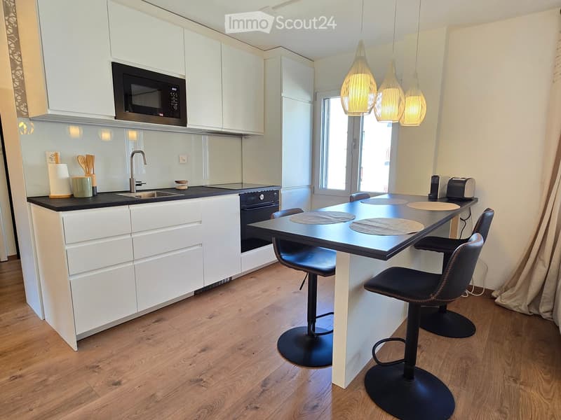 Modern kitchen with white cabinets, black countertops, a sink, oven, microwave, and a small dining area with two stools.