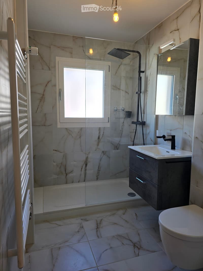 Modern bathroom with marble tiles, a glass-enclosed shower, a sink with a modern faucet, a toilet, and a radiator. The space is well-lit with natural light from a window.
