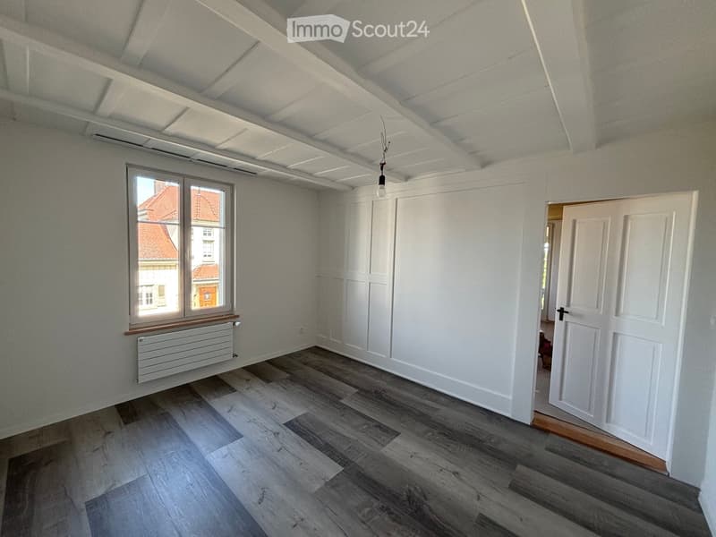 Empty room with wooden flooring, white walls, a window with a radiator, and a white door leading to another space.