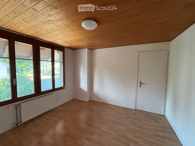 Empty room with wooden flooring, white walls, large windows, and a white door. The ceiling is wooden with a built-in light fixture.