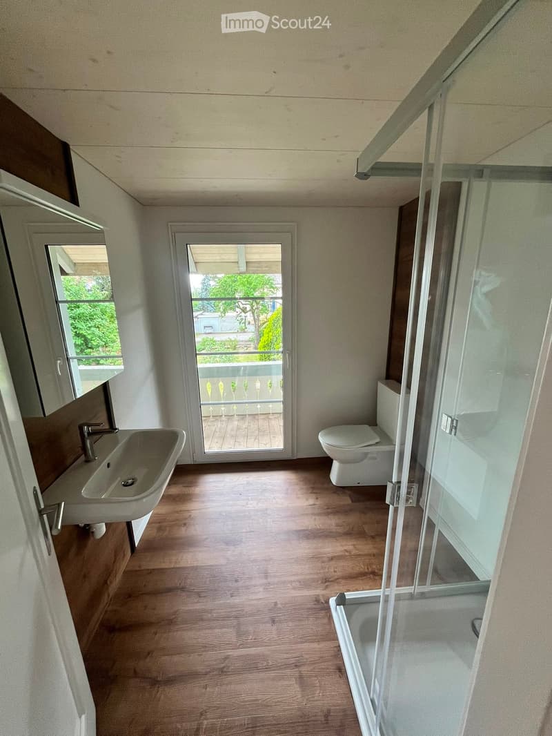 Modern bathroom with wooden flooring, a sink, a toilet, a shower, and a sliding glass door leading to a balcony.