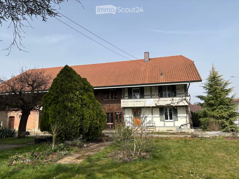 Two-story house with red-tiled roof, white and brown exterior, balcony, and surrounding garden with trees and shrubs.