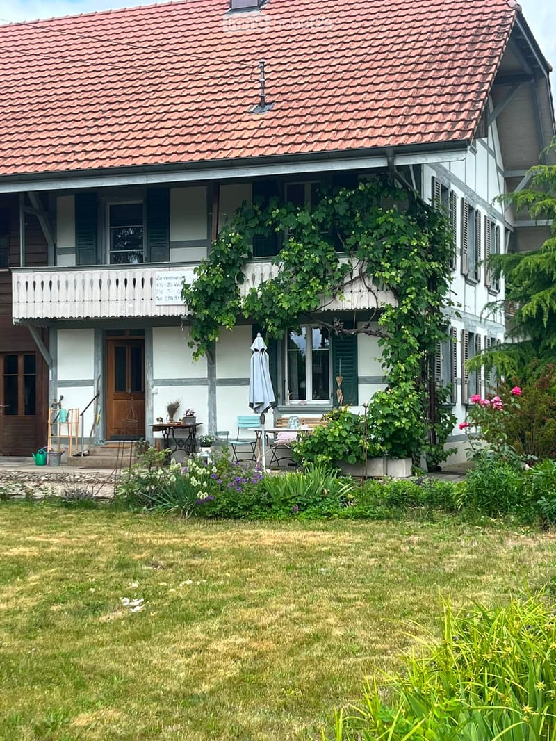 Two-story house with red-tiled roof, white walls, and green shutters. Features a balcony with a railing, a small garden with flowers, and outdoor furniture including a table and chairs.
