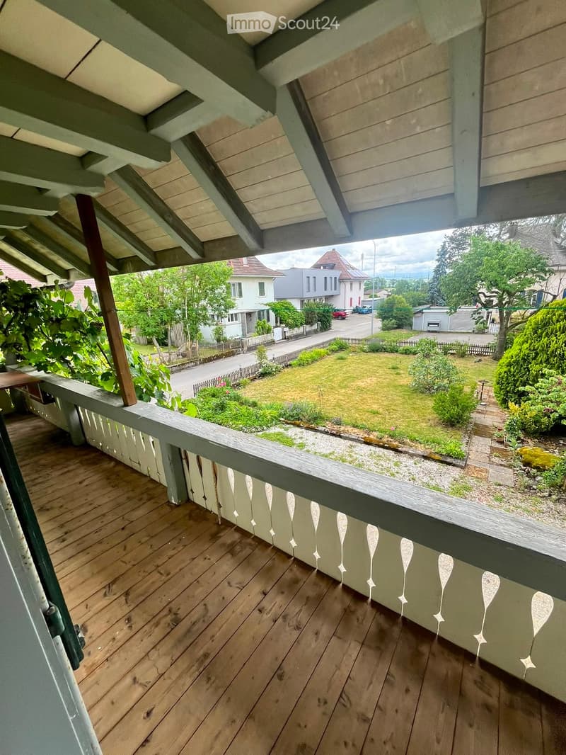 Wooden balcony with railing, overlooking a garden and street with houses.
