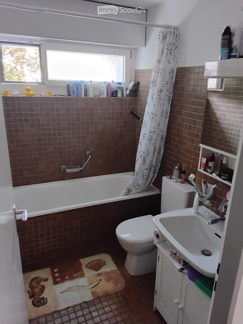 Bathroom with a bathtub, toilet, sink, and shower curtain. Brown tiled walls and floor, white fixtures, and a small window letting in natural light.