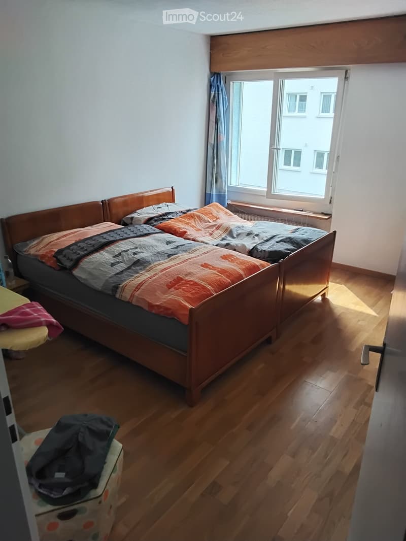 Bedroom with wooden floor, single bed with colorful quilt, window with curtain, radiator, and a small stool with items on it.