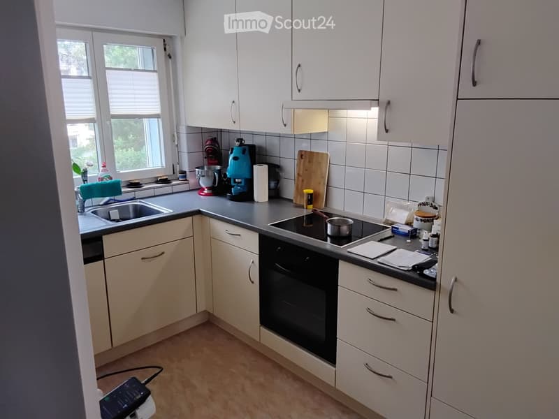 Modern kitchen with white cabinets, grey countertops, and various appliances including a sink, oven, coffee maker, and mixer. The space is well-lit with natural light from a window on the left side.