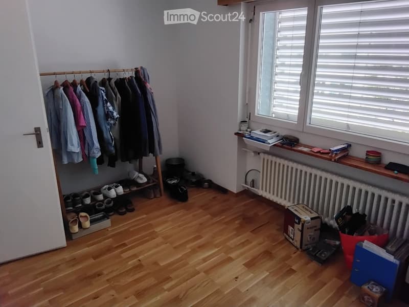 A small, well-lit room with wooden flooring, a clothing rack with hanging clothes, a shoe rack with several pairs of shoes, a radiator, and a shelf with various items including books and a small sink.
