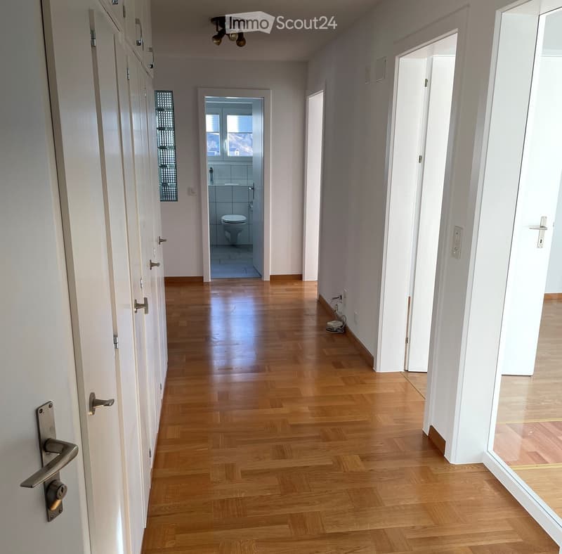 A long, narrow hallway with wooden flooring, white walls, and several doors on both sides. A small bathroom is visible at the end of the hallway.