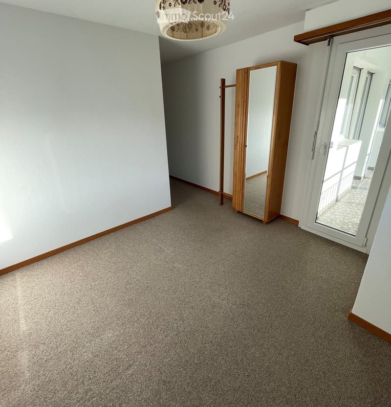 Empty room with light-colored walls, carpeted floor, and a large mirror on a wooden stand. A sliding glass door leads to a balcony area.