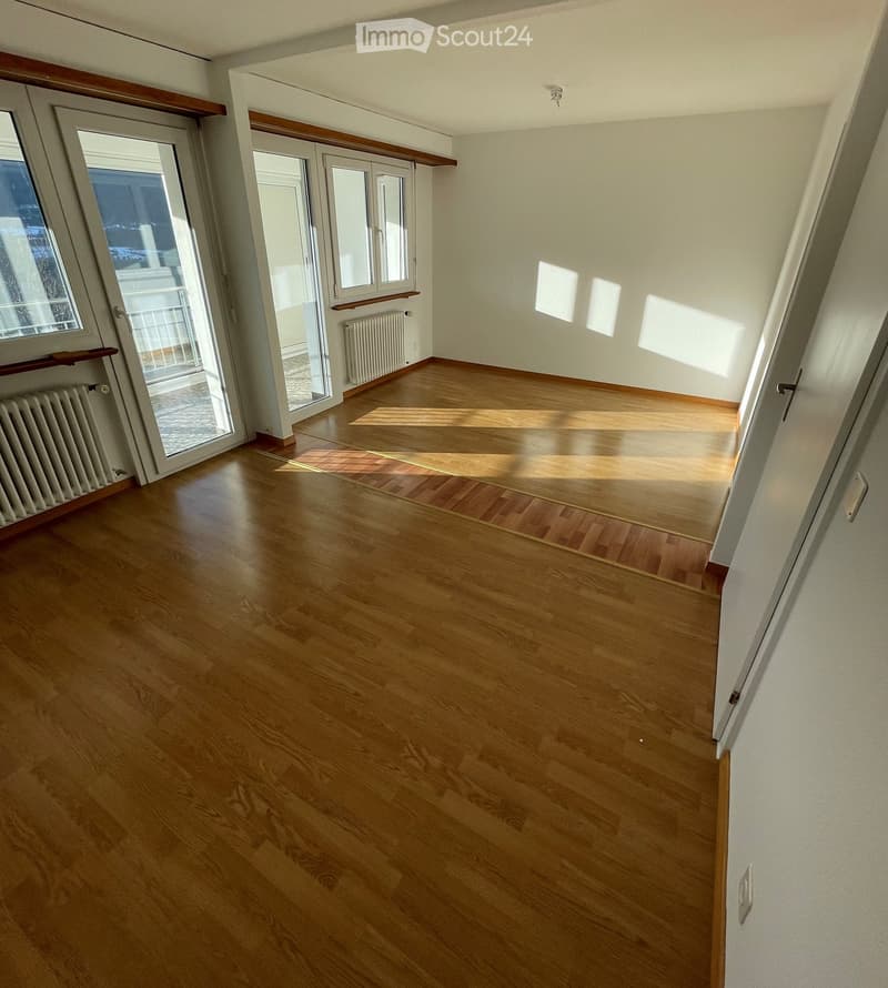 Empty room with wooden flooring, white walls, large windows, and a sliding glass door leading to a balcony.