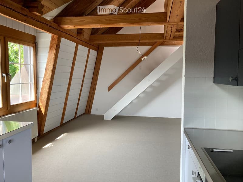 Unfurnished Room. Empty attic space with exposed wooden beams, white walls, and a large window letting in natural light. The floor is concrete, and there are no furnishings or decorations.