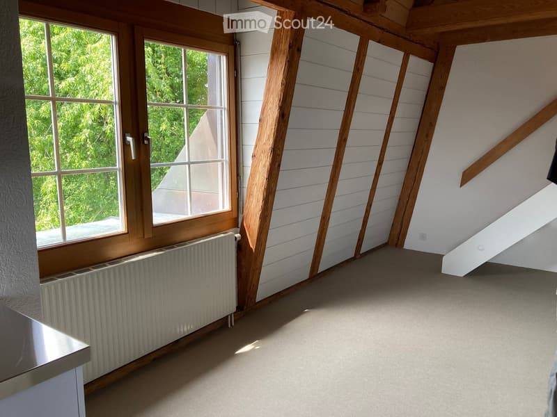 Unfurnished Room. Empty attic room with wooden beams, white paneled walls, large window, and radiator.