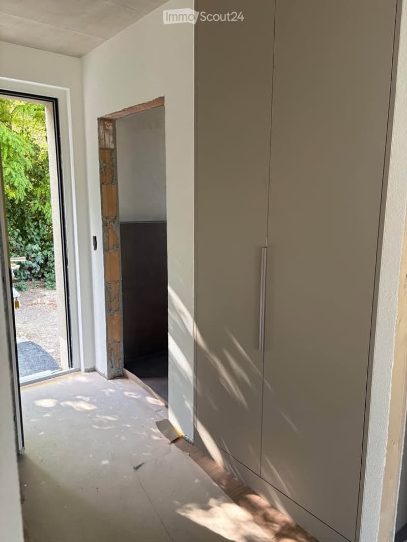 Empty room with white walls, tiled floor, large window with view of greenery, and a partially open doorway leading to another space.