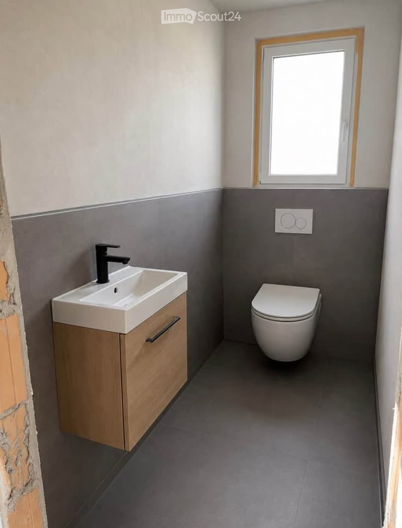 Modern bathroom with a white sink, black faucet, wooden cabinet, and white toilet. Grey tiled walls and floor, with a small window providing natural light.