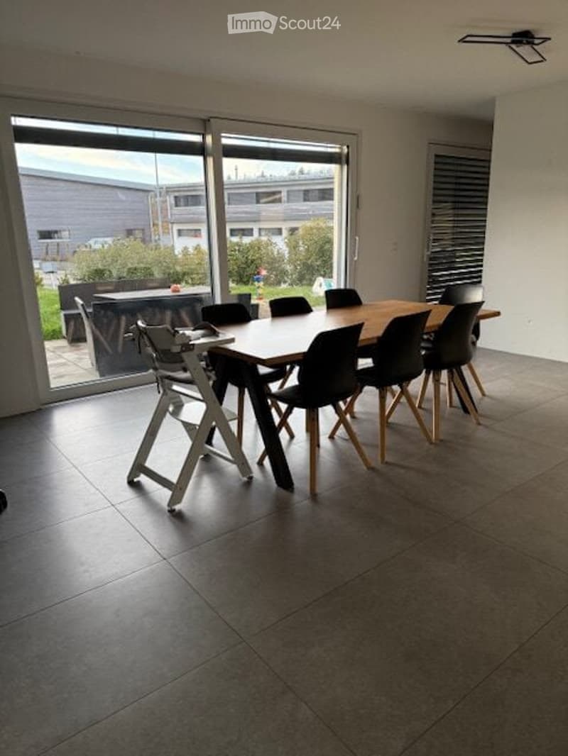 Bright dining area with large glass sliding doors leading to an outdoor space, featuring a wooden dining table surrounded by black chairs and a baby chair.