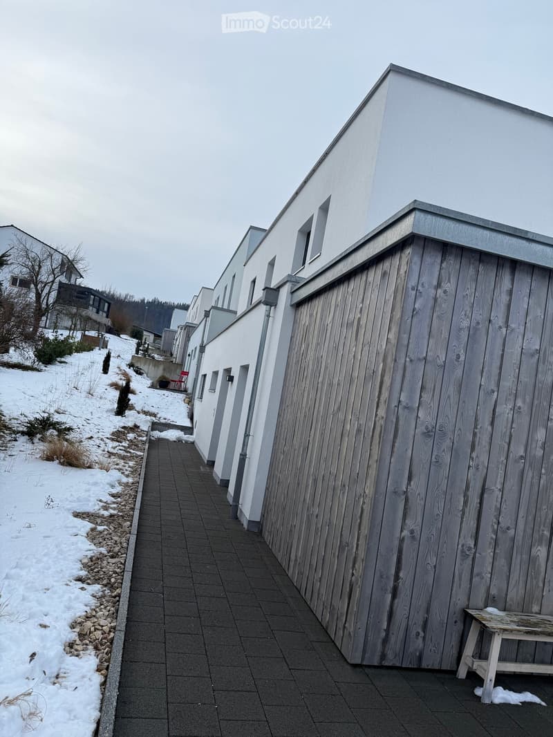 A snow-covered pathway leads between modern buildings with white and wooden facades, featuring a small wooden bench on the right side.