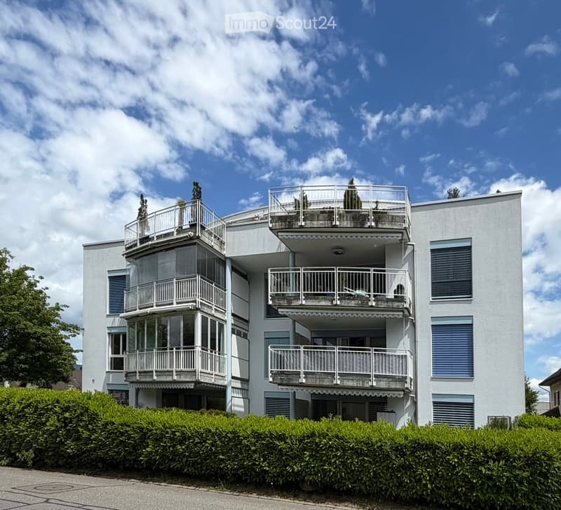 Modern multi-story building with multiple balconies and large windows, surrounded by greenery.