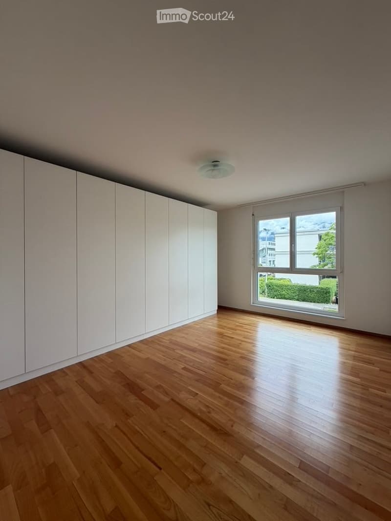 Empty room with wooden flooring, white built-in wardrobes, and large window with view of greenery.