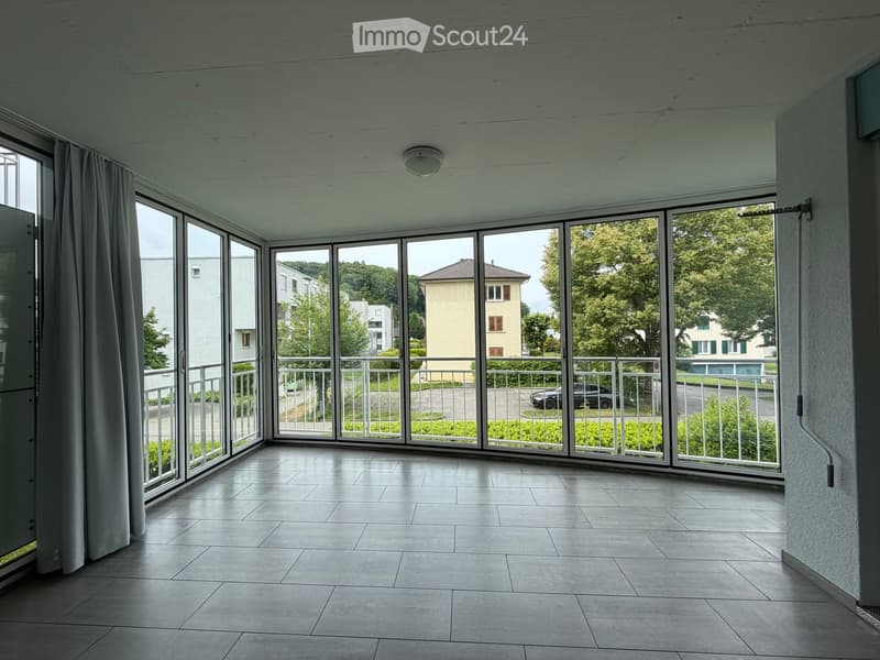 Empty room with large windows and a tiled floor, offering a view of the surrounding area and greenery.