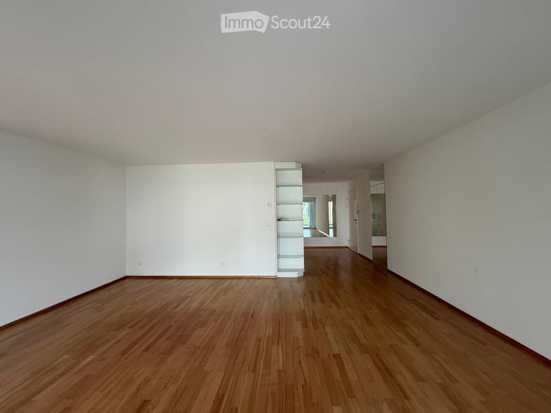 Empty room with wooden flooring and white walls, featuring a small glass shelf unit and a doorway leading to another room.