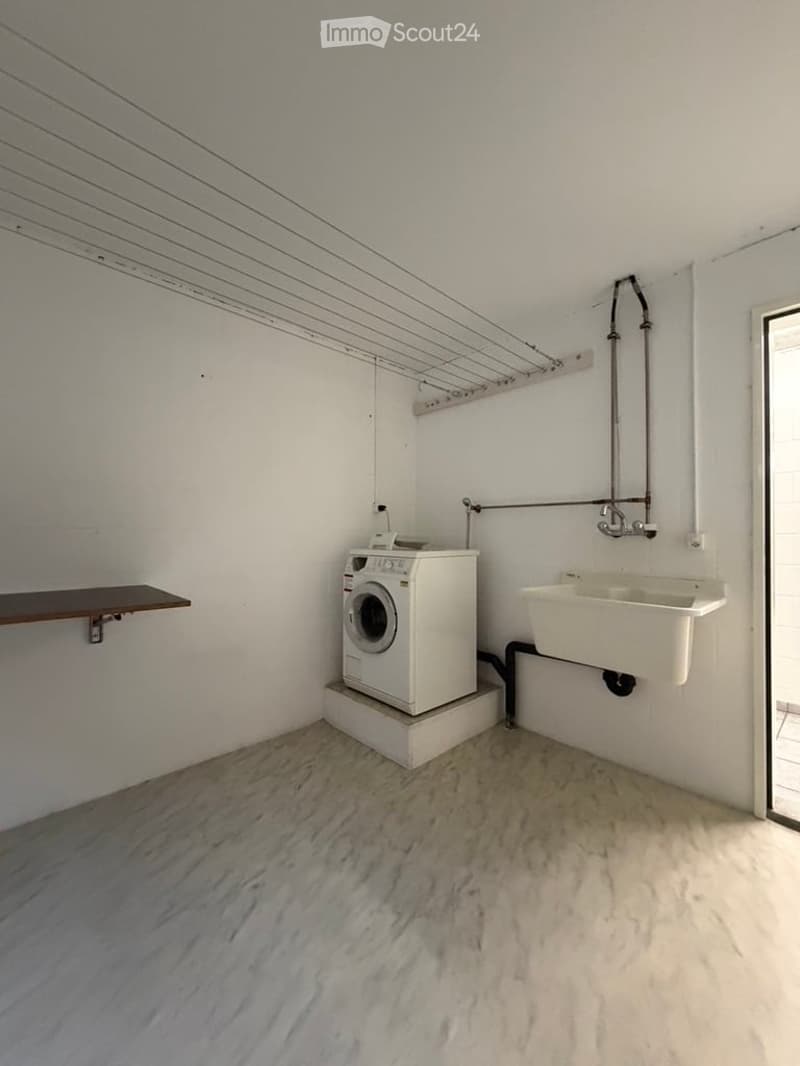 Bathroom with a white washing machine, a sink, and a wooden shelf. The floor is marble, and the walls are white with a metal rack above the washing machine.