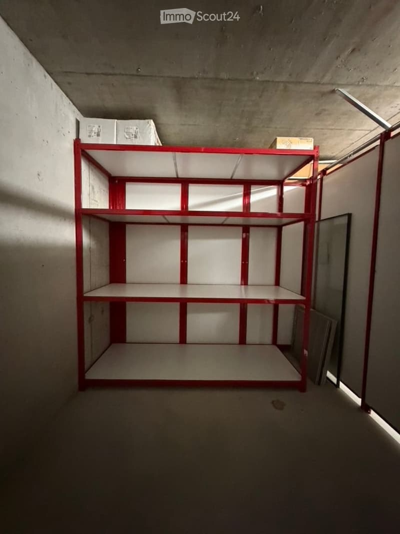 Empty room with concrete walls and floor, featuring a red metal shelving unit with multiple empty shelves.