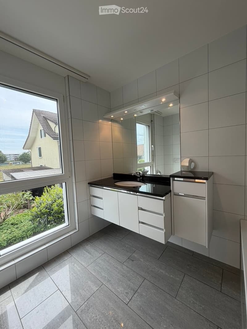 Modern bathroom with white tiled walls, grey tiled floor, black countertop, white cabinets, and a large mirror with integrated lighting.