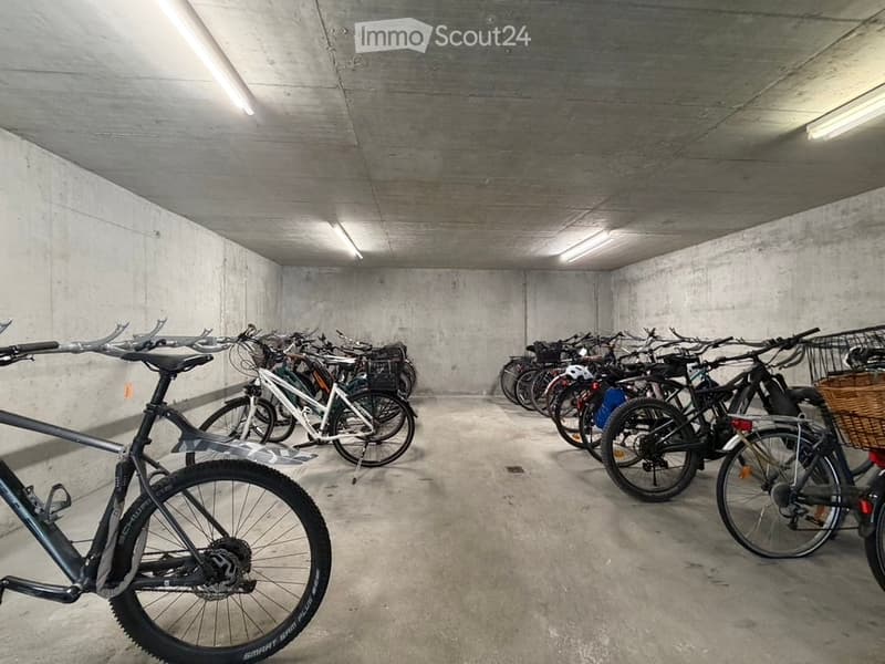 Underground parking area with multiple bicycles lined up against the wall, concrete walls and ceiling, fluorescent lighting.