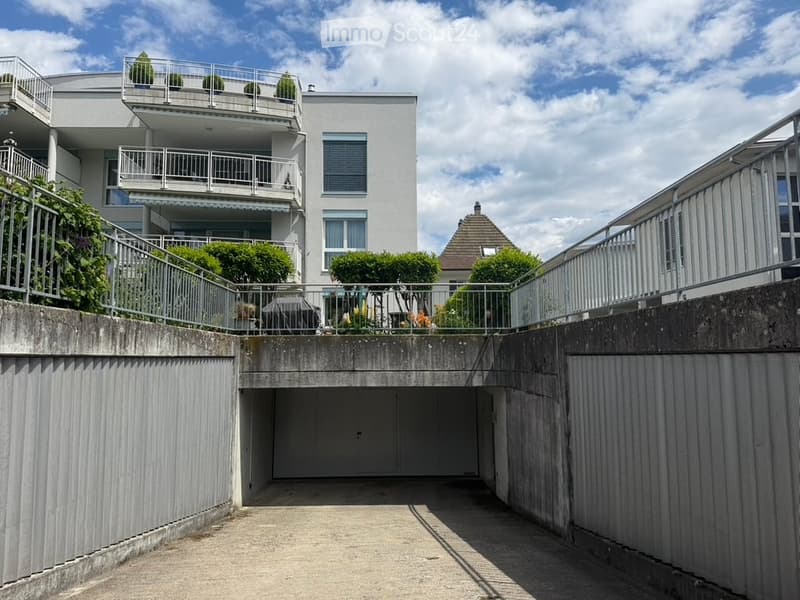 Modern multi-story building with balconies, garage entrance, and surrounding greenery.