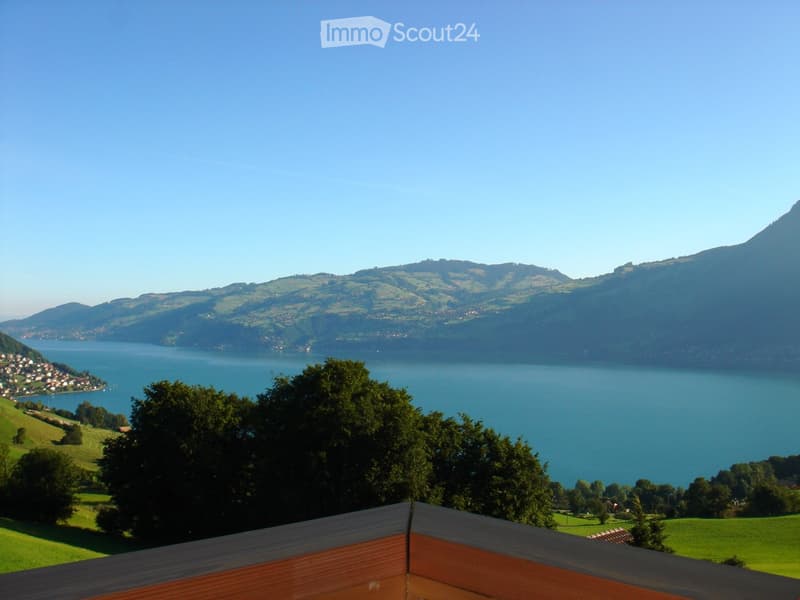 Balcony overlooking a serene lake surrounded by green hills and distant mountains under a clear blue sky.