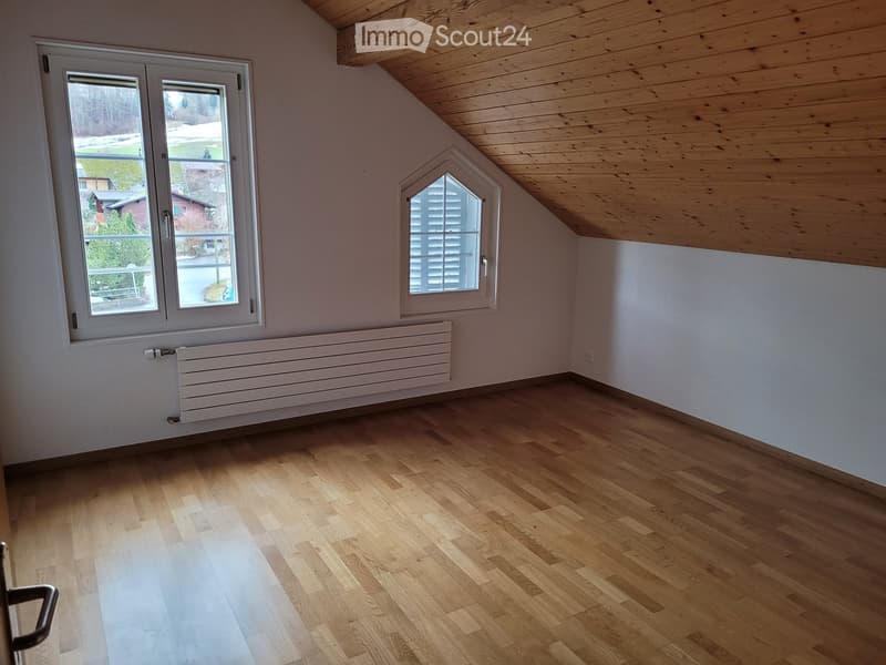 Empty attic room with wooden flooring, white walls, and exposed wooden ceiling beams. Features a large window with a view and a smaller dormer window, along with a white radiator below the main window.