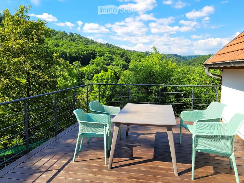 Balcony with wooden decking, metal railing, and a table with four chairs overlooking a lush green forest.