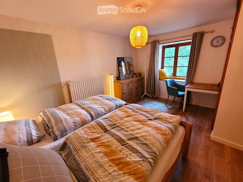 Cozy bedroom with a large double bed, patterned bedding, a wooden dresser with a mirror, a desk with a chair, and a window with curtains. The room features warm lighting from a hanging lamp and a floor lamp, and has wooden flooring.